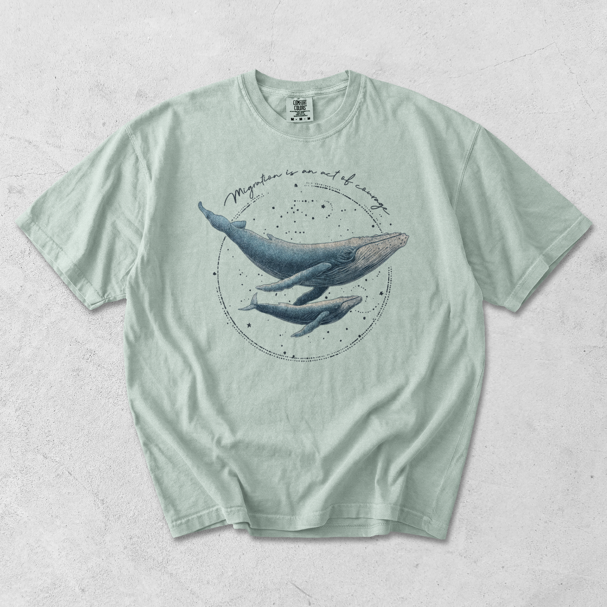 Mint green t-shirt with whale graphic on a textured surface, with writing migration is and act of courage